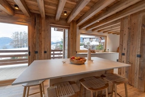 A wooden interior with a large table and bar stools, a window with a view of mountains, and a basket of fruit on the table. - Chalet Combloux, Megève, Mont-Blanc View (Combloux)