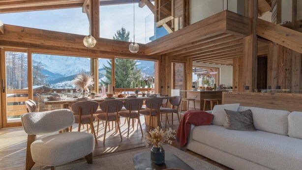 Ampla sala de estar com uma grande janela que oferece vista para uma paisagem montanhosa coberta de neve. Há uma área de jantar com mobiliário de madeira e uma cozinha visível através da janela.
