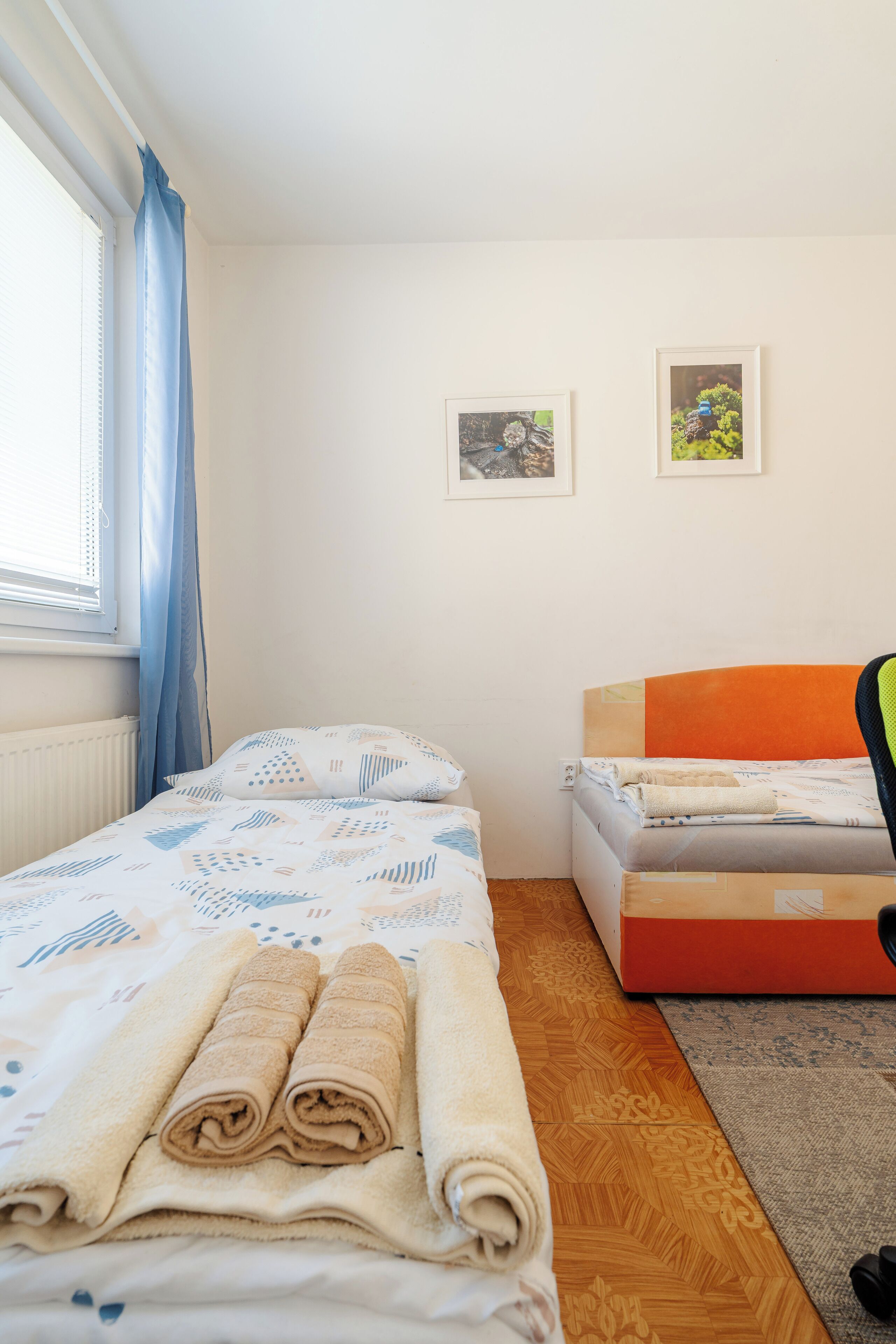 A bedroom with two beds, a window with blue curtains, and two framed pictures on the wall.