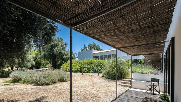 Un espacio al aire libre techado con terraza de madera, zona de estar con banco y sillas, y vista a un edificio moderno al fondo.