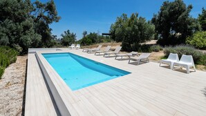 Piscina rectangular con cubierta de madera, rodeada de sillas reclinables blancas y vegetación.