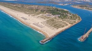 Vista aérea de una zona costera con playa de arena, una pequeña península con edificios y un muelle que se adentra en el agua.