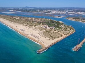 An aerial view of a coastal area with a sandy beach, a small peninsula with buildings, and a pier extending into the water. - Olea Green Studio 1, Tavira, Algarve (Querença)