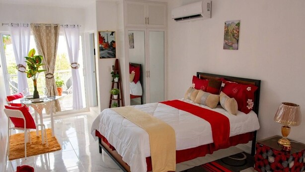 A bedroom with a bed, a bedside table, a chair, a small table, and a window with curtains. - Room-royal Terrace Queen 1 (Puerto Plata)