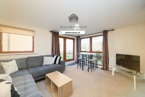 A modern living room with a grey sofa, a wooden coffee table, a dining table with chairs, a TV, and large windows with curtains. - 2 bed flat with parking (West Sussex)