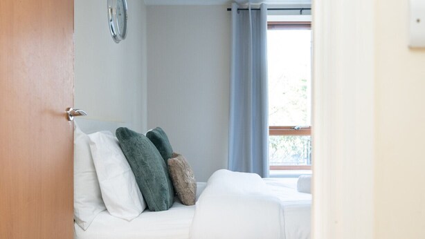 Um quarto com cama, almofadas, janela com cortinas e uma porta.