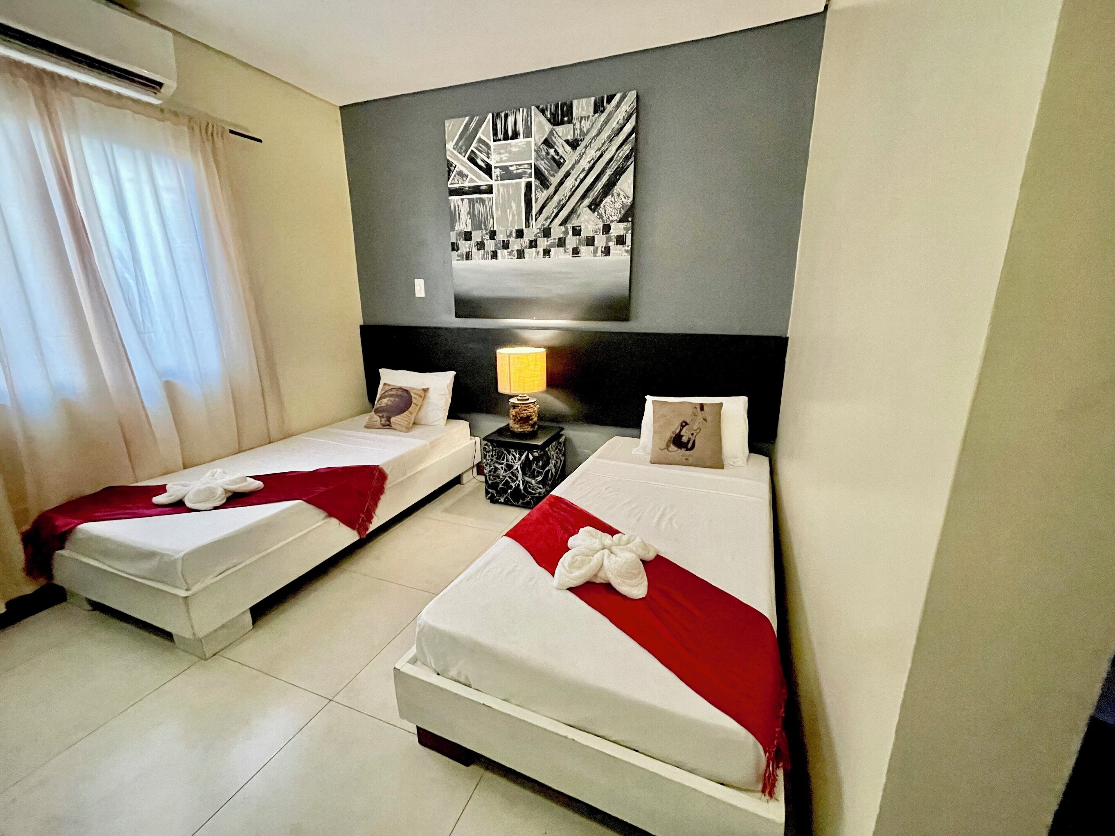 Habitación de hotel con dos camas individuales, una mesita de noche con lámpara, televisor montado en la pared y un cuadro abstracto grande.