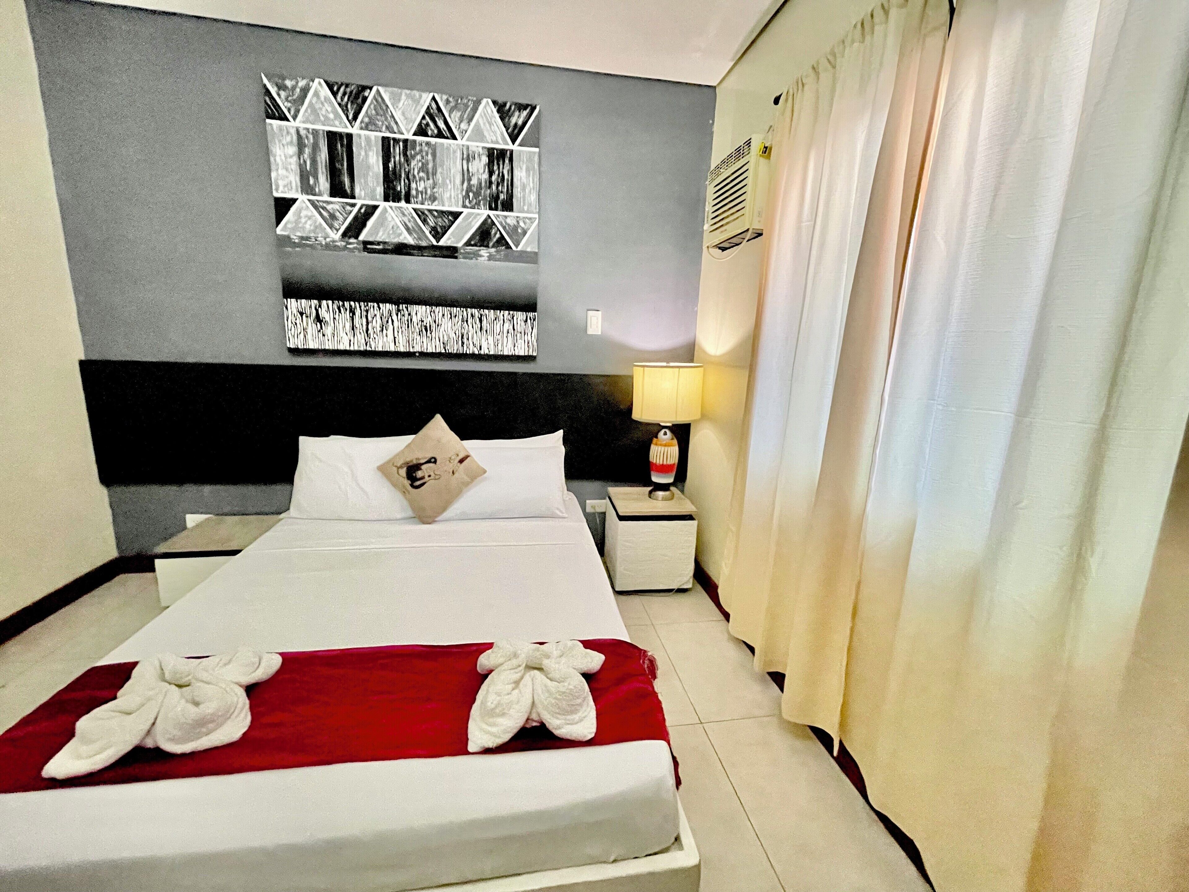 A hotel room with a neatly made bed, a wall-mounted air conditioner, and a bedside lamp.