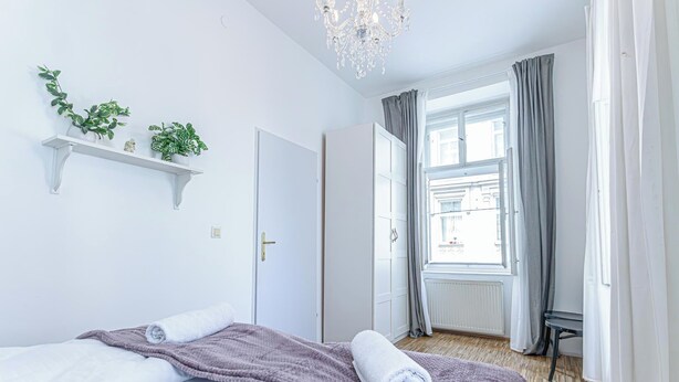 A hotel room with a bed, a window with curtains, a chandelier, and a plant shelf. - Elegant Spacious 2BR City Center Stay (Vienna)