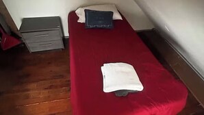 Una habitació petita amb un llit individual, un estovat vermell, un coixí blanc i un coixí blau fosc. Hi ha un petit armari gris al mur i una fotografia emmarcada a sobre. La habitació té un paviment de fusta i un sostre inclinat.