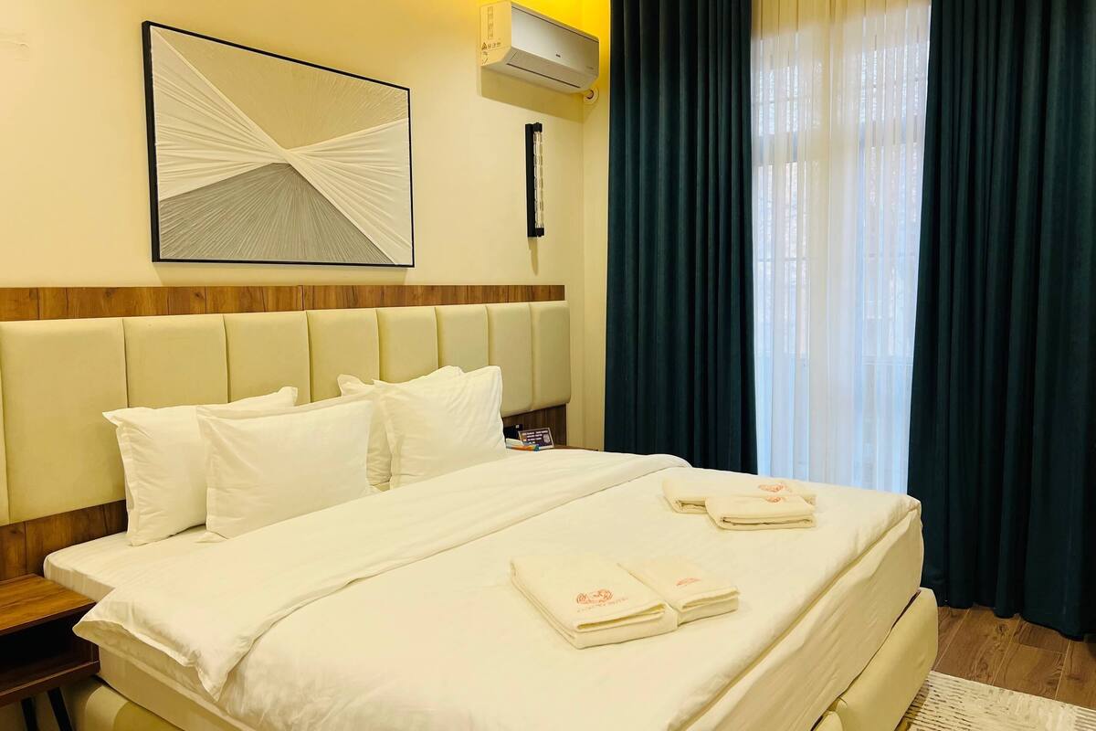 Een hotelkamer met een bed, wit beddengoed, een hoofdbord, een wandairconditioner, een lijst met een kunstwerk en een raam met donkere gordijnen.