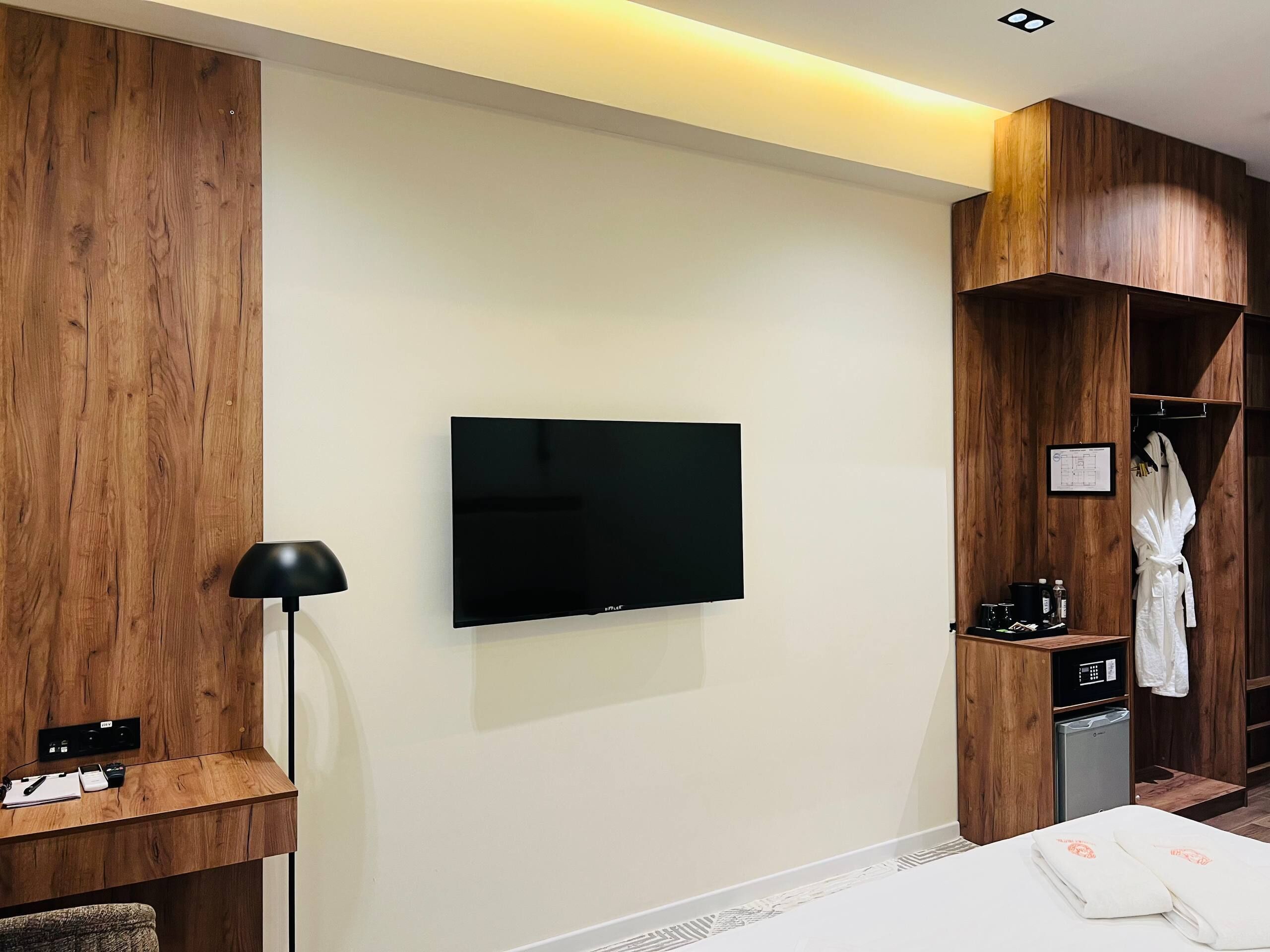 Una habitación de hotel moderna con una televisión de pantalla plana en la pared, un escritorio con lámpara, un mini refrigerador y una bata colgada en un perchero.