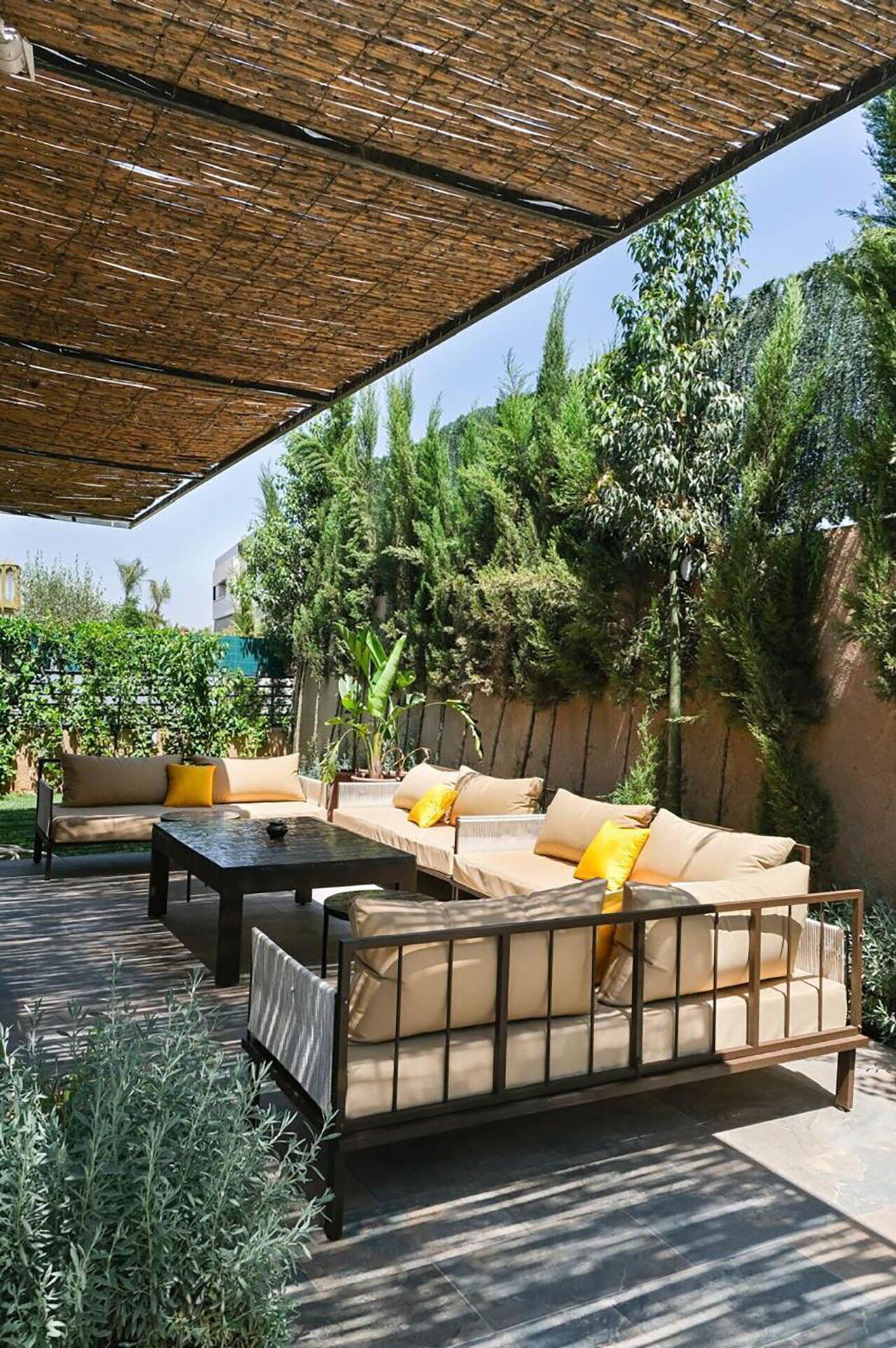 Un área de estar al aire libre con un sofá y una mesa, rodeada de follaje y una pérgola.