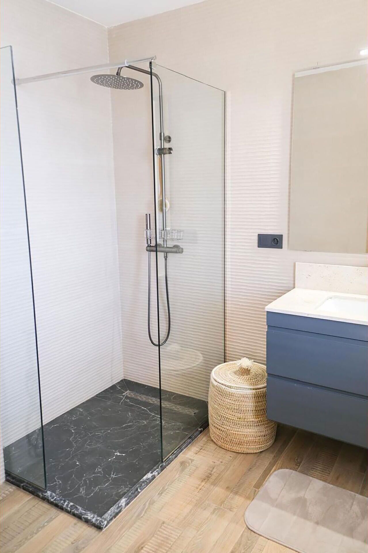 Een moderne badkamer met een douchecabine van glas, een houten vloer, een blauwe wastafelmeubel en een gevlochten mand.