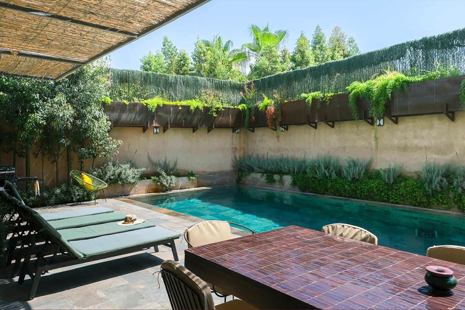 Un área de piscina al aire libre con una mesa y sillas, rodeada de follaje y un espacio de descanso cubierto.