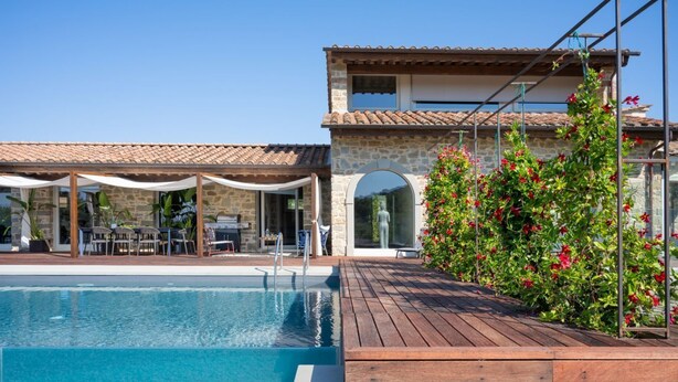 Un área de piscina con terraza de madera, muros de piedra y una zona de estar cubierta.
