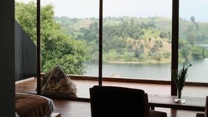 Habitación con ventanas amplias que dan a un lago y un bosque. Se observa una cama, una silla y una mesita con una planta.