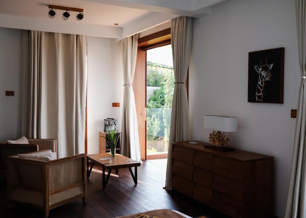 Una habitación con sofá de madera, una mesita con un florero, un mueble de madera y un ventanal con cortinas.