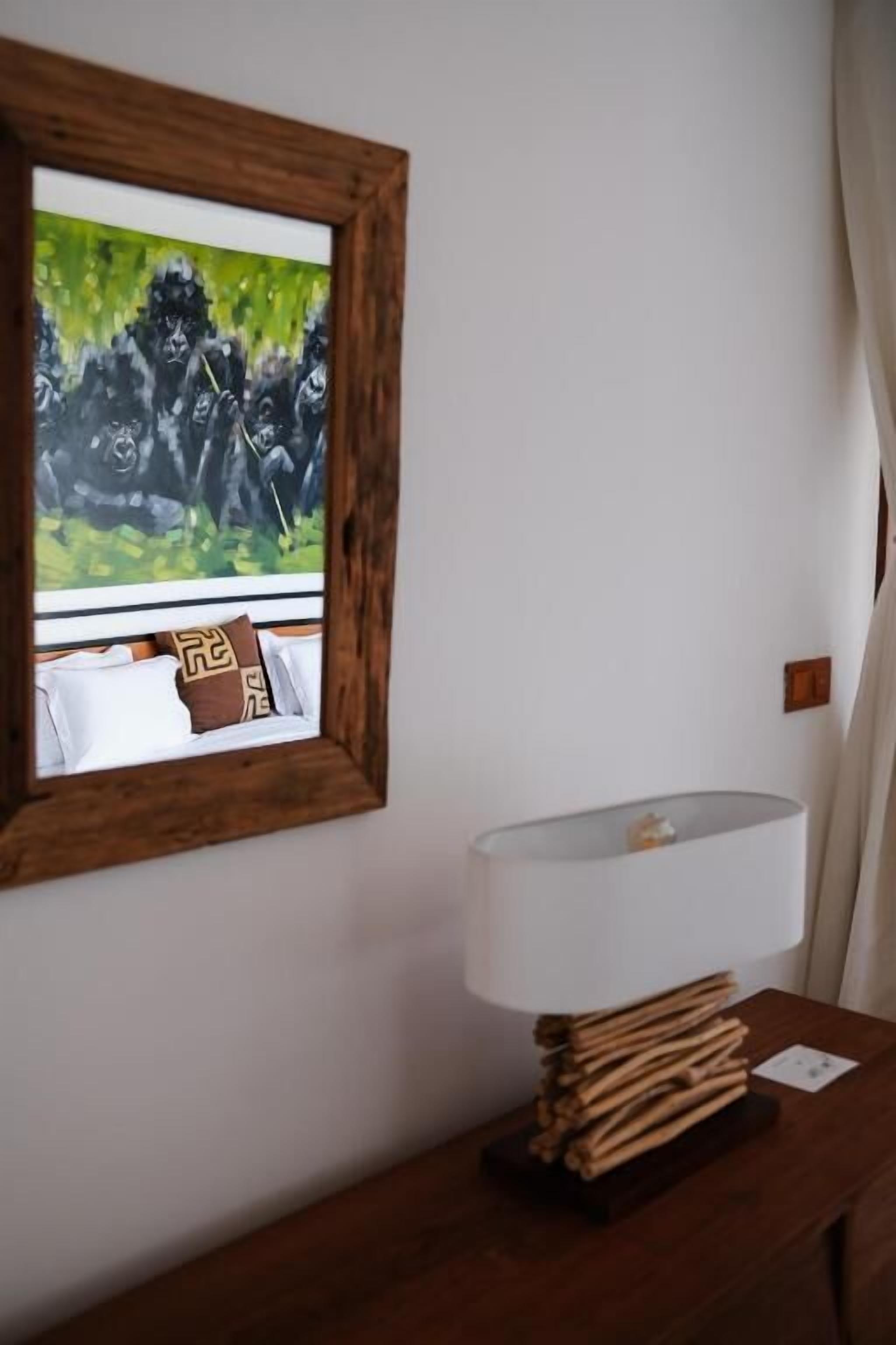 A room with a wooden bedside table, a white lamp, a framed painting of gorillas, and a bed with white linens and patterned pillows.