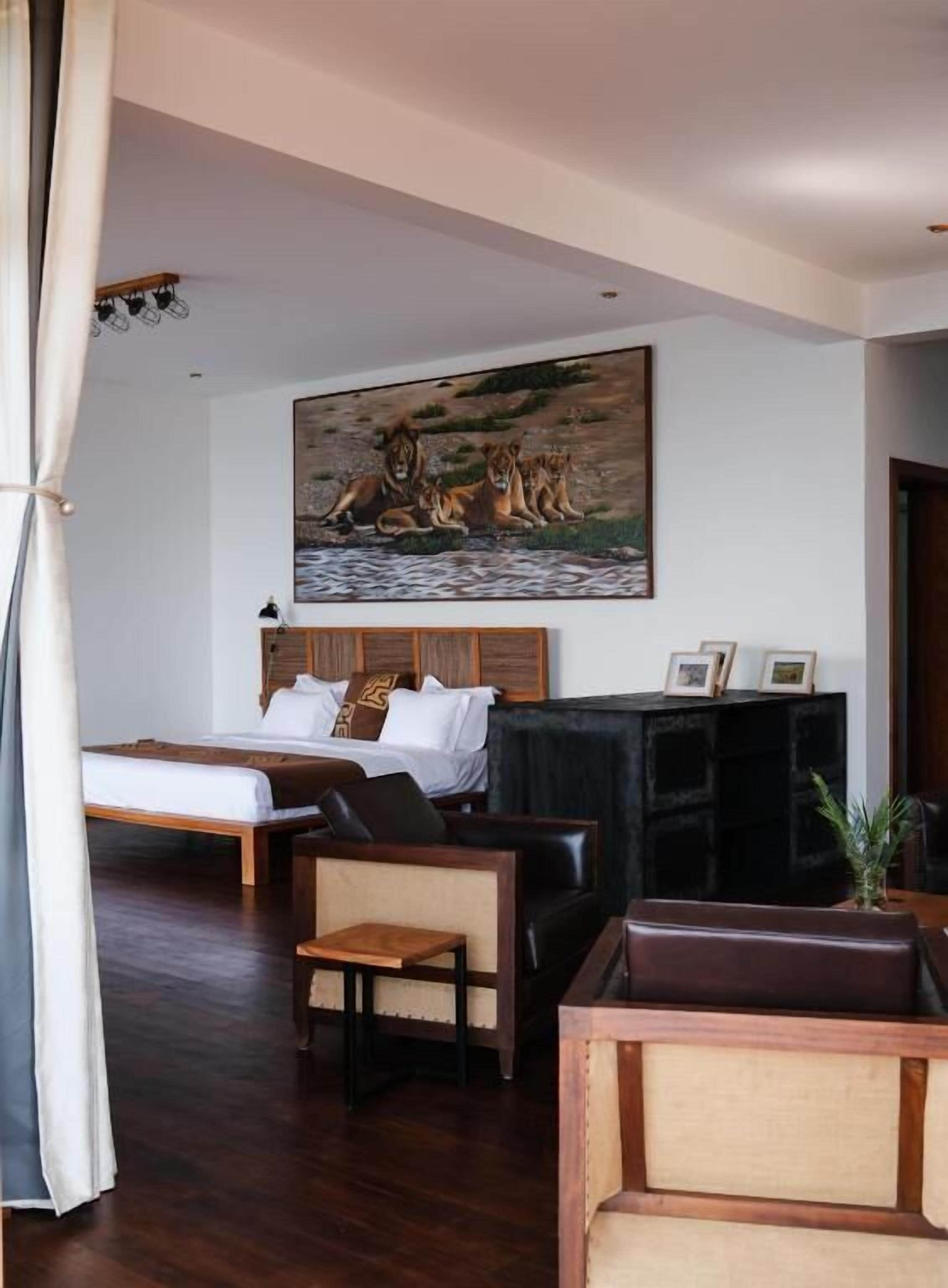 Une chambre d’hôtel avec un lit, un tableau représentant des lions, un meuble noir pour la télévision et d’autres appareils, et une table basse en bois.