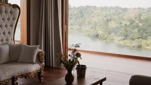 Habitación con un ventanal grande que da a un área boscosa. Hay un sofá capitonado con detalles ornamentales, una mesa de centro de madera con un jarrón de flores y una pequeña mesa con una silla.