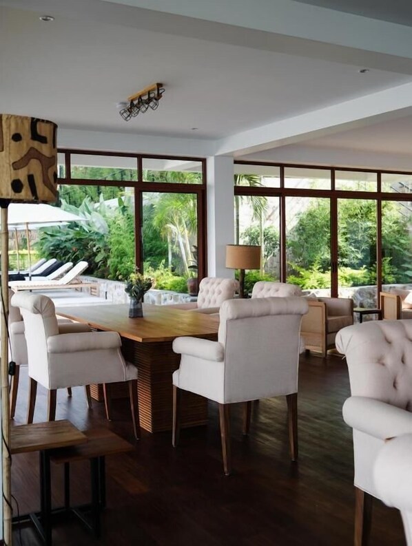 A spacious room with a large wooden dining table surrounded by white upholstered chairs, a sofa, and a lamp. Large windows provide a view of lush greenery outside. - Umurobyi Lodge (Bwishyura)
