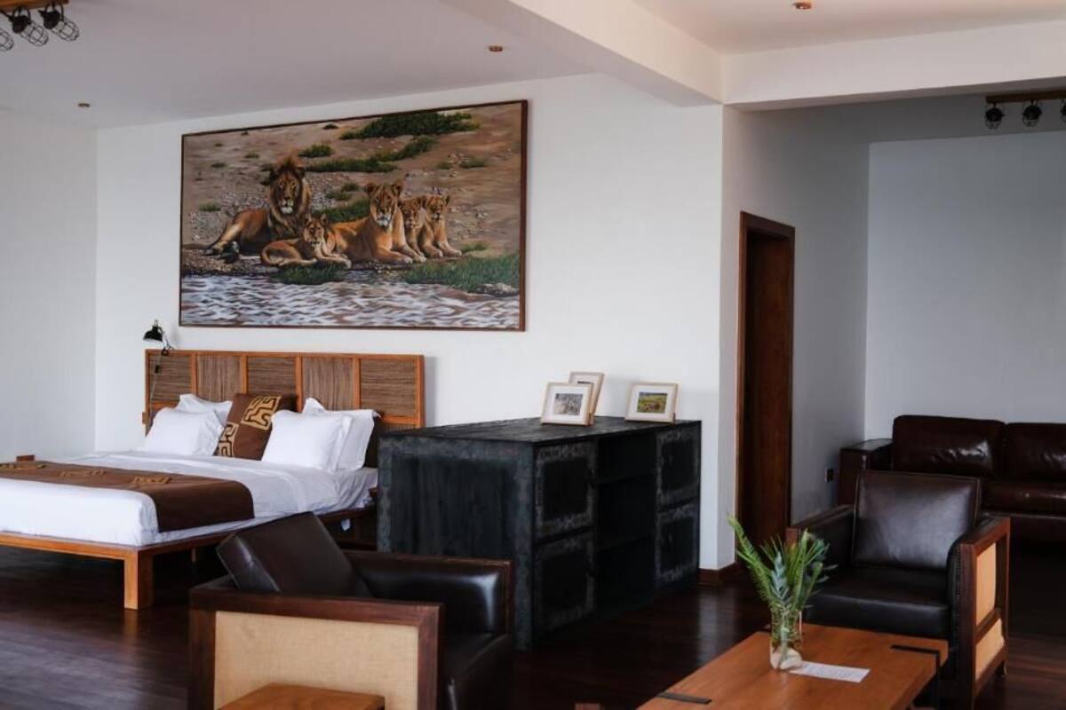 Une chambre d’hôtel avec un grand lit, une tête de lit en bois, une console noire ornée de photos encadrées, un canapé en cuir marron et un tableau représentant des lions près d’un point d’eau.