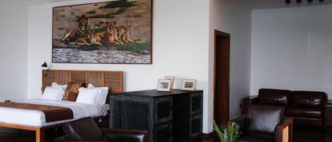 A hotel room with a large bed, a wooden headboard, a black console table with framed photos, a brown leather sofa, and a painting of lions by a waterhole.