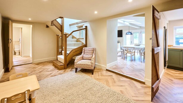 A spacious room with a herringbone floor, a wooden staircase, a patterned armchair, and a dining area visible through an open door. - Host & Stay | The Coral Cottage (Morley Saint Botolph)