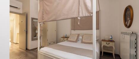 A four-poster bed with a canopy, a bedside table with a lamp, a framed picture on the wall, and a radiator.