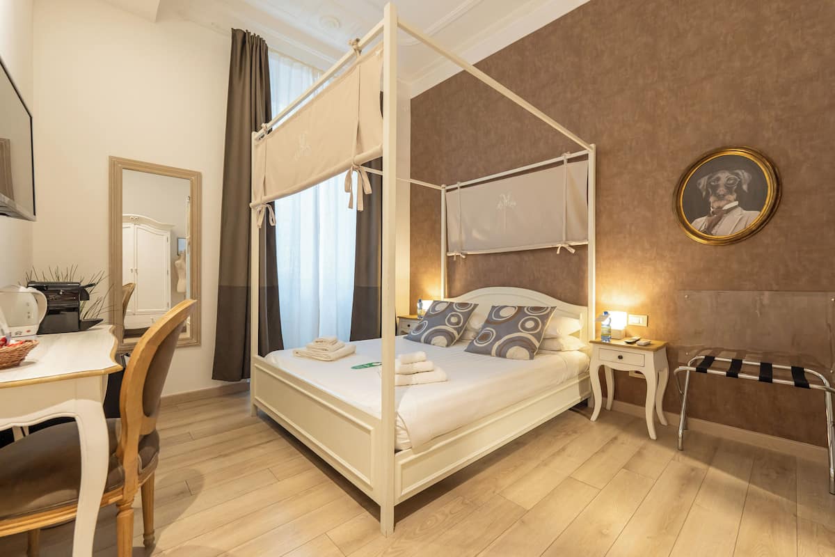 A four-poster bed with a canopy, a bedside table with a lamp, a chair, a desk with a kettle, and a framed picture on the wall.