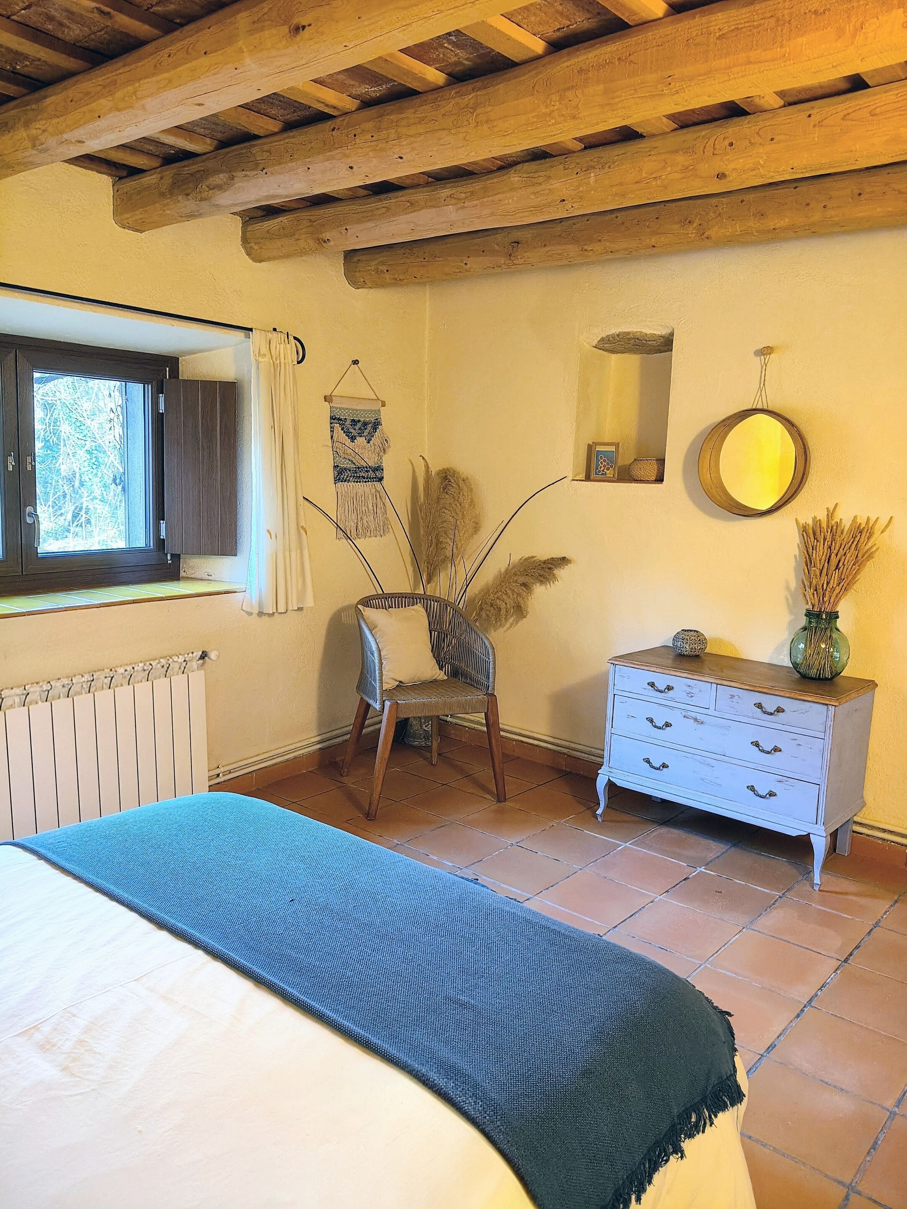 Una habitación con cama, una silla, cómoda, ventana y espejo.