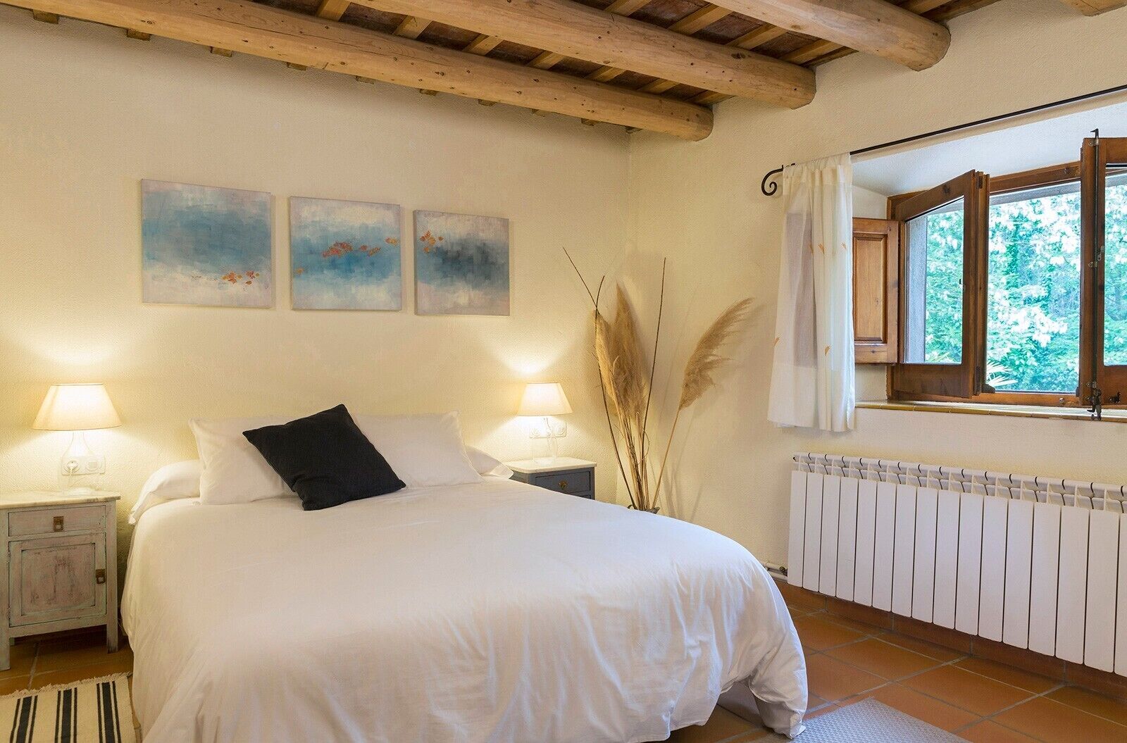 Un dormitorio con una cama, dos mesitas de noche, un ventanal con cortinas y tres cuadros colgados en la pared.