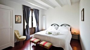 Een slaapkamer met een bed, een stoel, een bankje, een raam met gordijnen en een nachtkastje met een lamp.