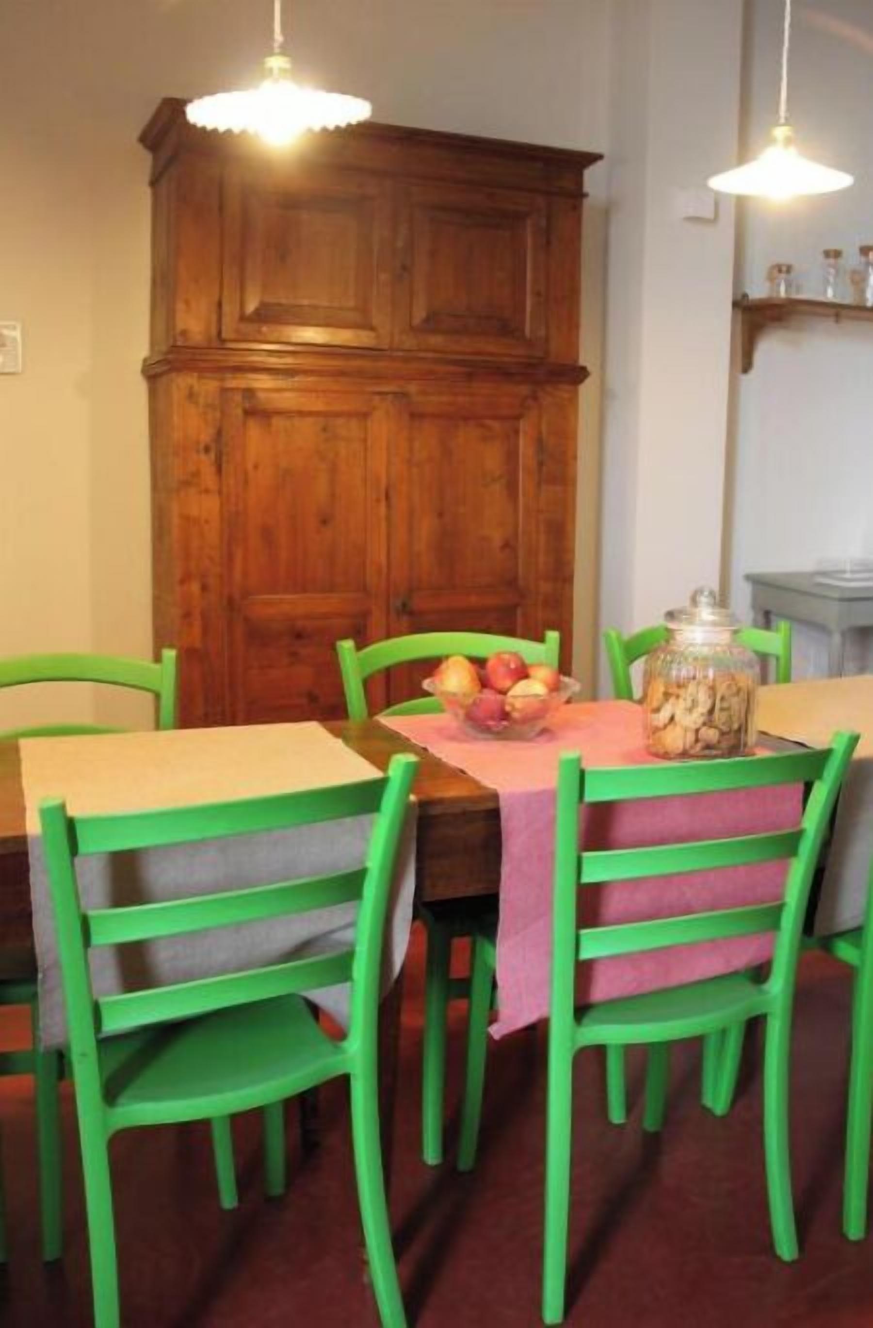 Un coin repas avec un buffet en bois, des chaises vertes, une table recouverte d’une nappe rose et un panier de fruits.