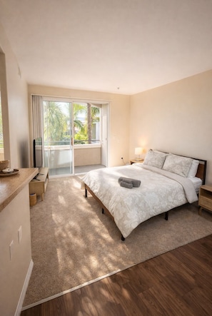 A bedroom with a large bed, a nightstand, a lamp, and a view of a balcony with palm trees. - Sunny Marina Del Rey 1BA Getaway (Marina del Rey)