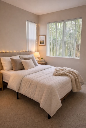 A bedroom with a large bed, a nightstand, a lamp, a picture on the wall, and a window with a view of palm trees. - Sunny Marina Del Rey 1BA Getaway (Marina del Rey)