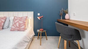 Ein Schlafzimmer mit Bett, Nachttisch, Lampe, Stuhl und Schreibtisch an einer blauen Wand.
