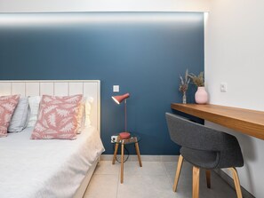 Quarto com cama, mesa de cabeceira, luminária, cadeira e escrivaninha em frente a uma parede azul.