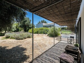 A covered outdoor area with a wooden deck, a seating area with a bench and chairs, and a view of a modern building in the background. - Olea Brown Studio 3, Tavira, Algarve (Querença)