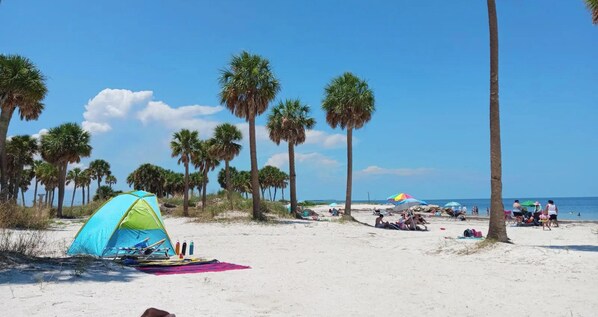 A beach with tents and umbrellas, palm trees, and people relaxing. - Family Coastal Home w/ EV Charger, Fenced Yard, Fire Pit & BBQ (Holiday)