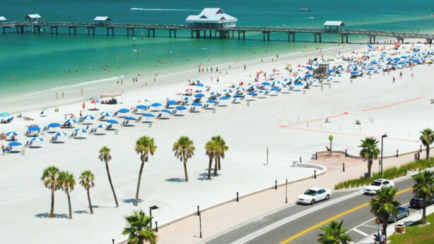 Een strand met rijen parasols en palmbomen, een pier die het water in loopt, en een weg ernaast.