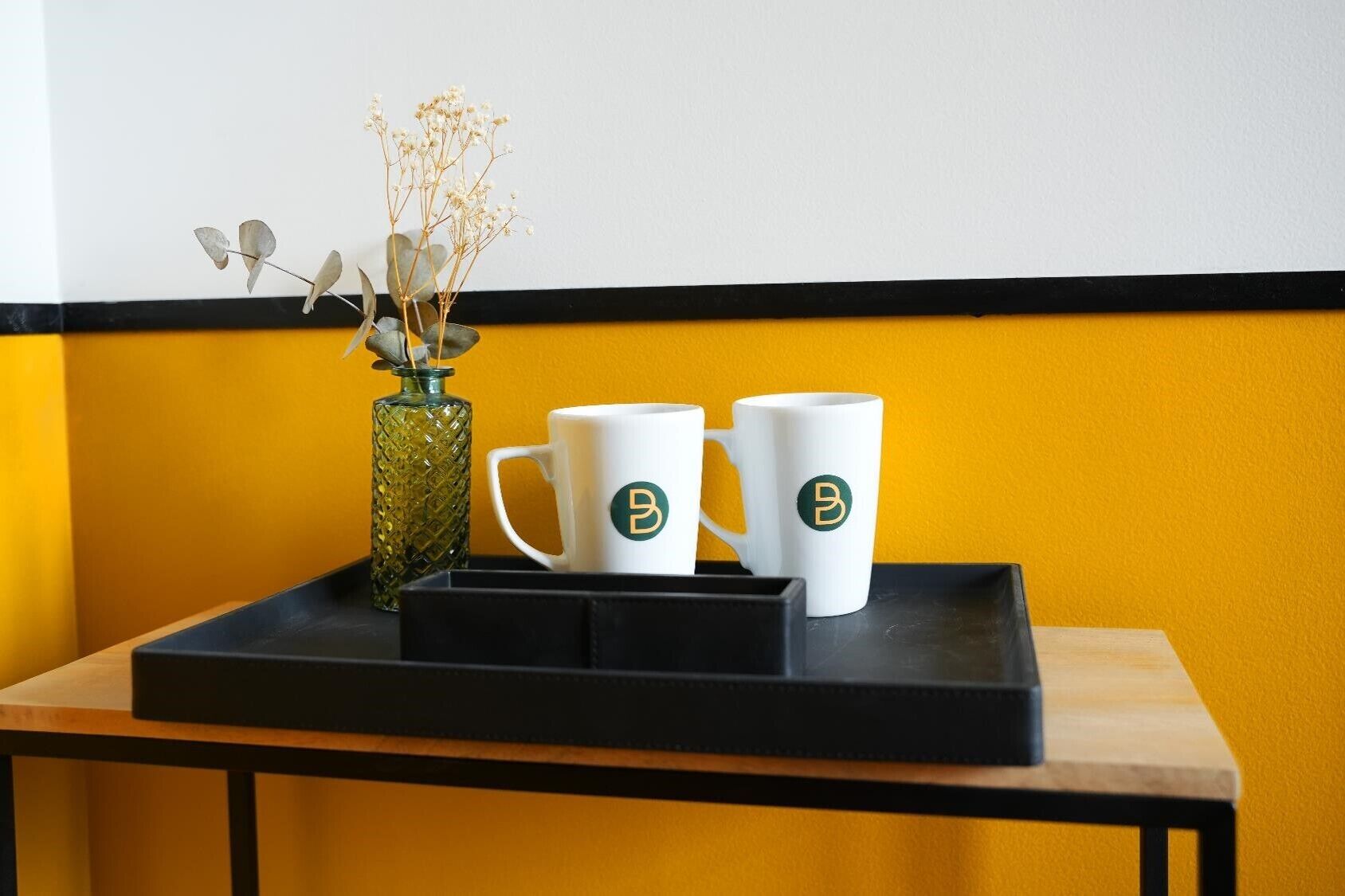 Een tafel met twee witte mokken met een logo, een glazen vaas met gedroogde bloemen, en een zwart dienblad.