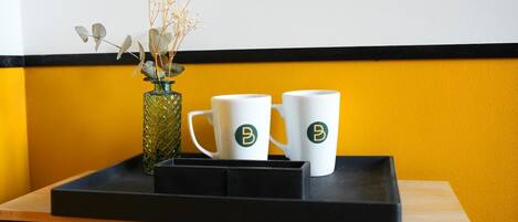 A table with two white mugs with a logo, a glass vase with dried flowers, and a black tray.