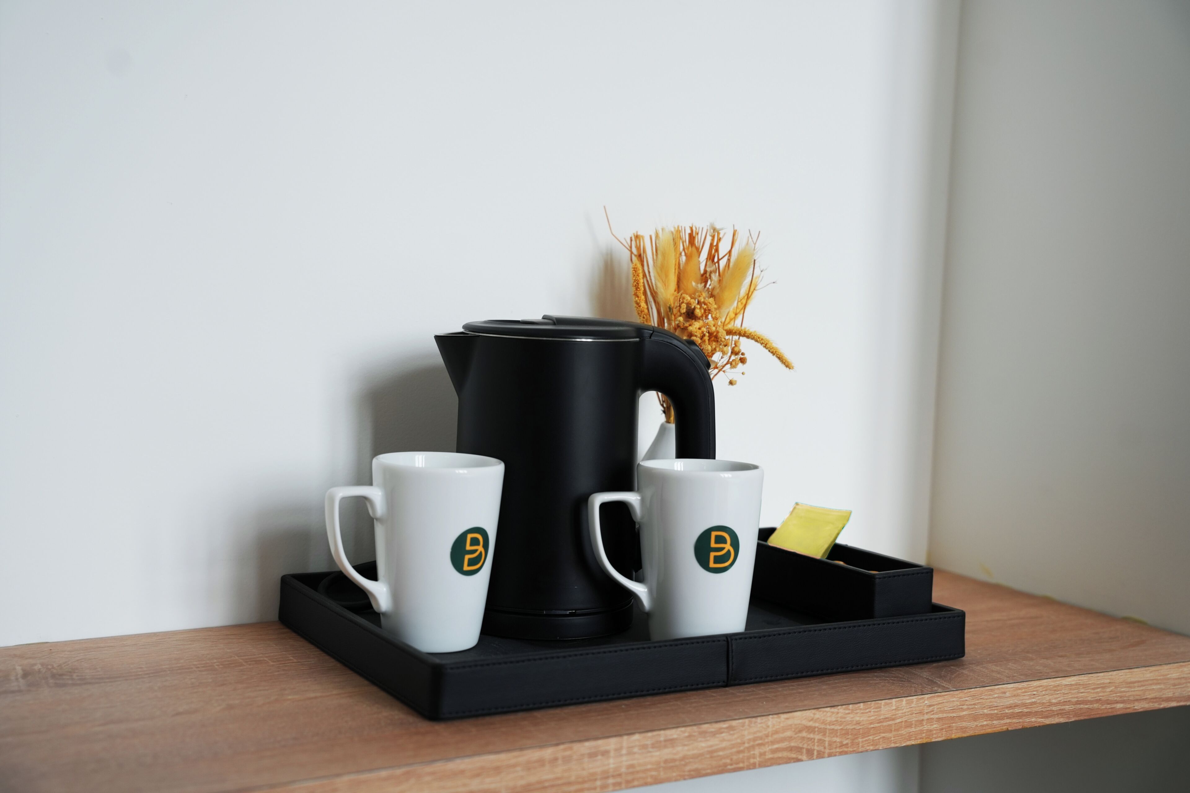 A black coffee maker, two white mugs with a logo, and a tray on a wooden shelf.