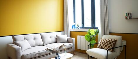 A living room with a sofa, armchair, and coffee table, a window with curtains, and a radiator.