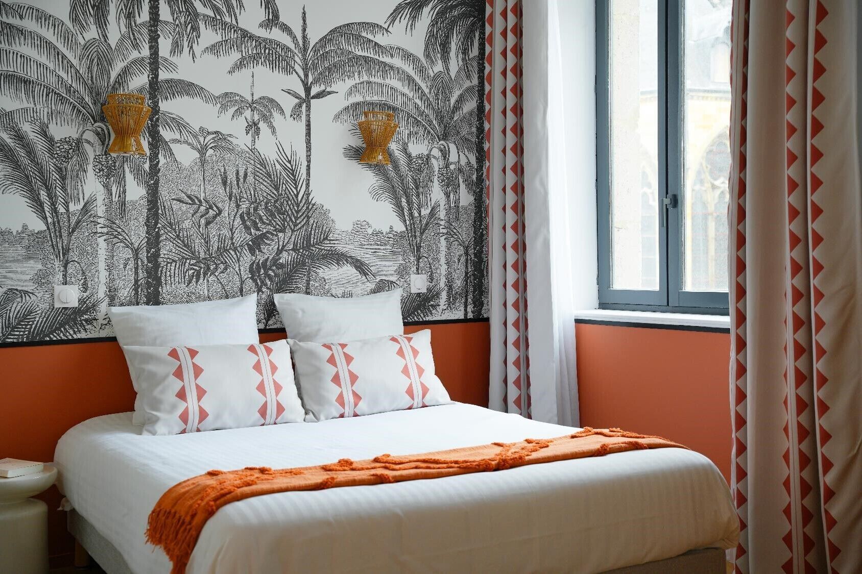 Una cama con ropa de cama blanca y detalles naranja, una pared con estampado de planta tropical, y una ventana con cortinas.