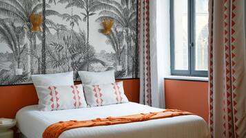 Una cama con ropa de cama blanca y detalles naranja, una pared con estampado de planta tropical, y una ventana con cortinas.