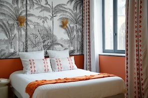Una cama con ropa de cama blanca y detalles naranja, una pared con estampado de planta tropical, y una ventana con cortinas.