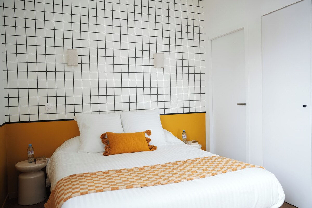 A neatly made bed with a checkered blanket, a bedside table with a bottle, and a wall with a grid pattern.